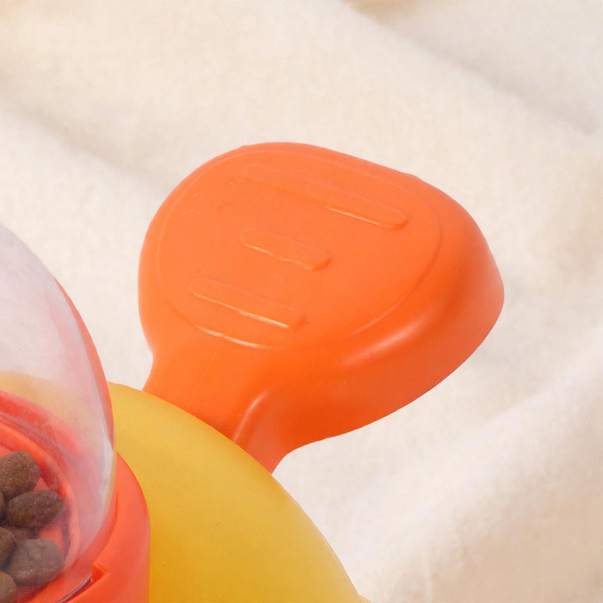 Close-up of the orange press pedal on the yellow duck treat dispenser, with the kibble-filled transparent dome partially visible.
