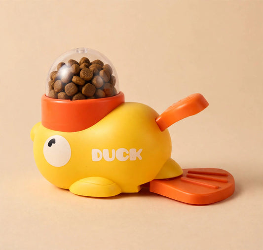 Yellow duck-shaped treat dispenser with a transparent dome full of kibble, an orange twist lid, an orange tail pedal, and DUCK printed on its side.