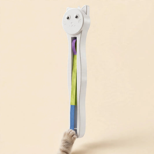 White cat-face-shaped wall-mount cat teaser with a long flat woven strap in purple, yellow-green, and blue segments, as a cat's paw reaches up to touch it.