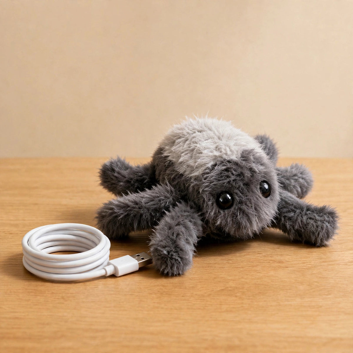 Gray plush spider toy lying on a wooden table next to a coiled white USB charging cable.