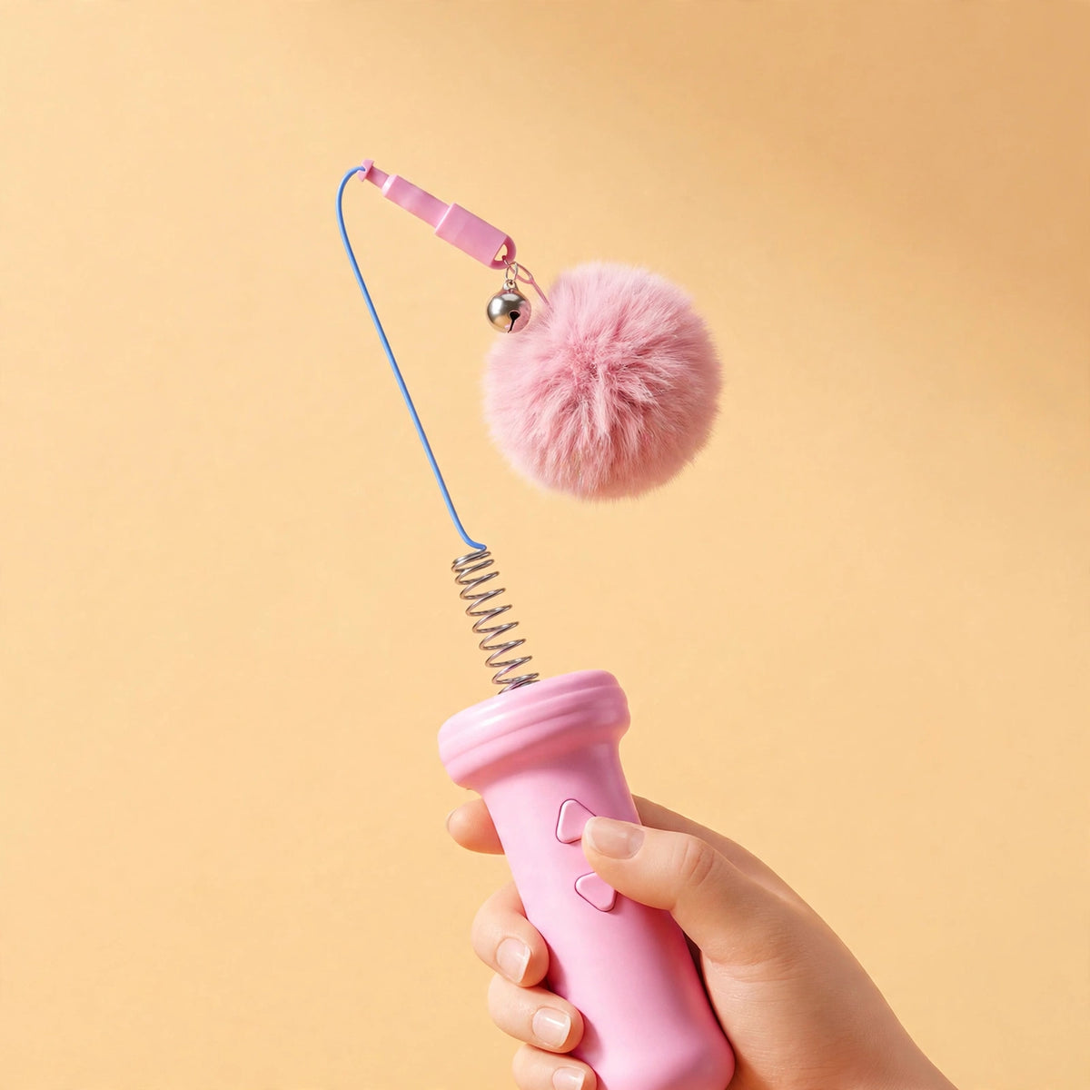 A hand pressing the retract button on the pink teaser wand as its spring coil, blue cord, and pink pom-pom ball head extend upward.
